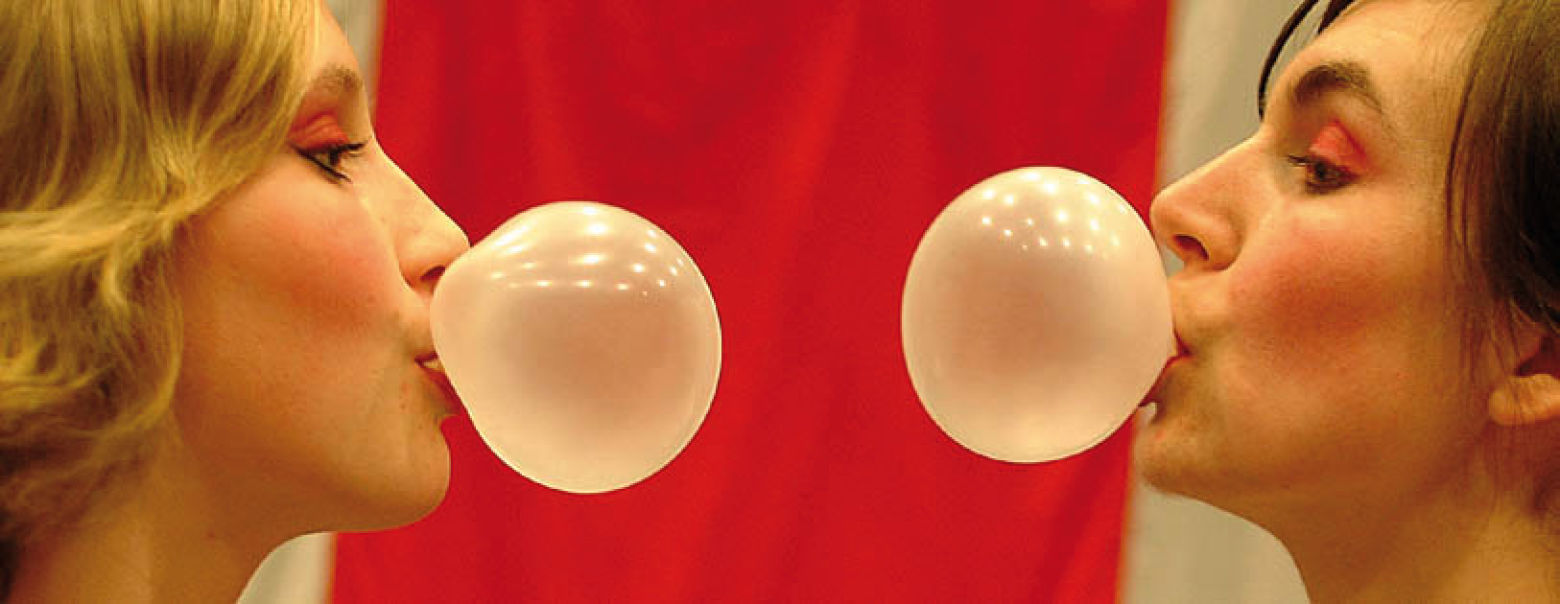 To kvinner i profil blåser tyggegummiboble mot hverandre. Det er rød bakgrunn.